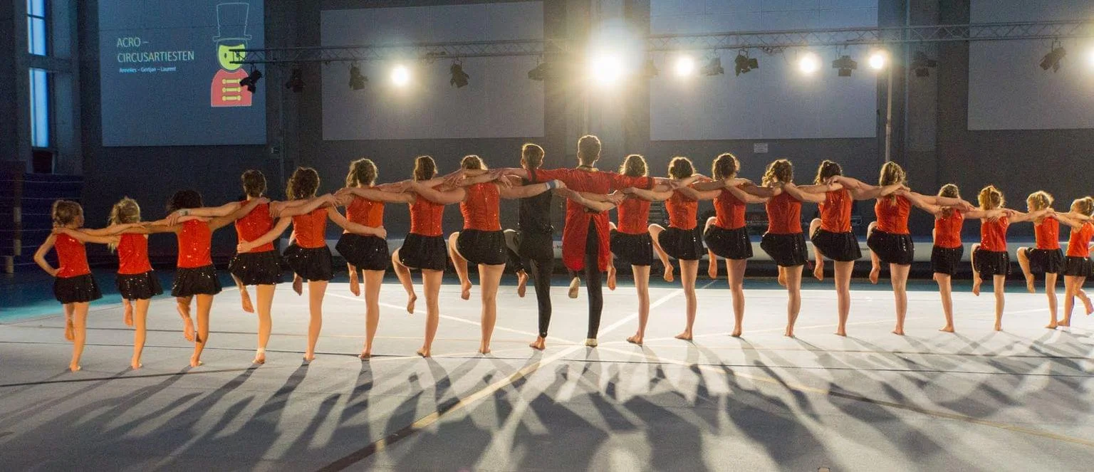Een groep jonge turners van Turnclub Olympia Zwijnaarde (TOZ) voert een prachtige, synchroon uitgevoerde teamroutine uit tijdens een turnshow. De atleten, gekleed in rode glinsterende tops en zwarte rokjes, staan schouder aan schouder en tillen hun benen in perfecte harmonie. Op de achtergrond verlichten theaterlampen de goed georganiseerde sporthal.
