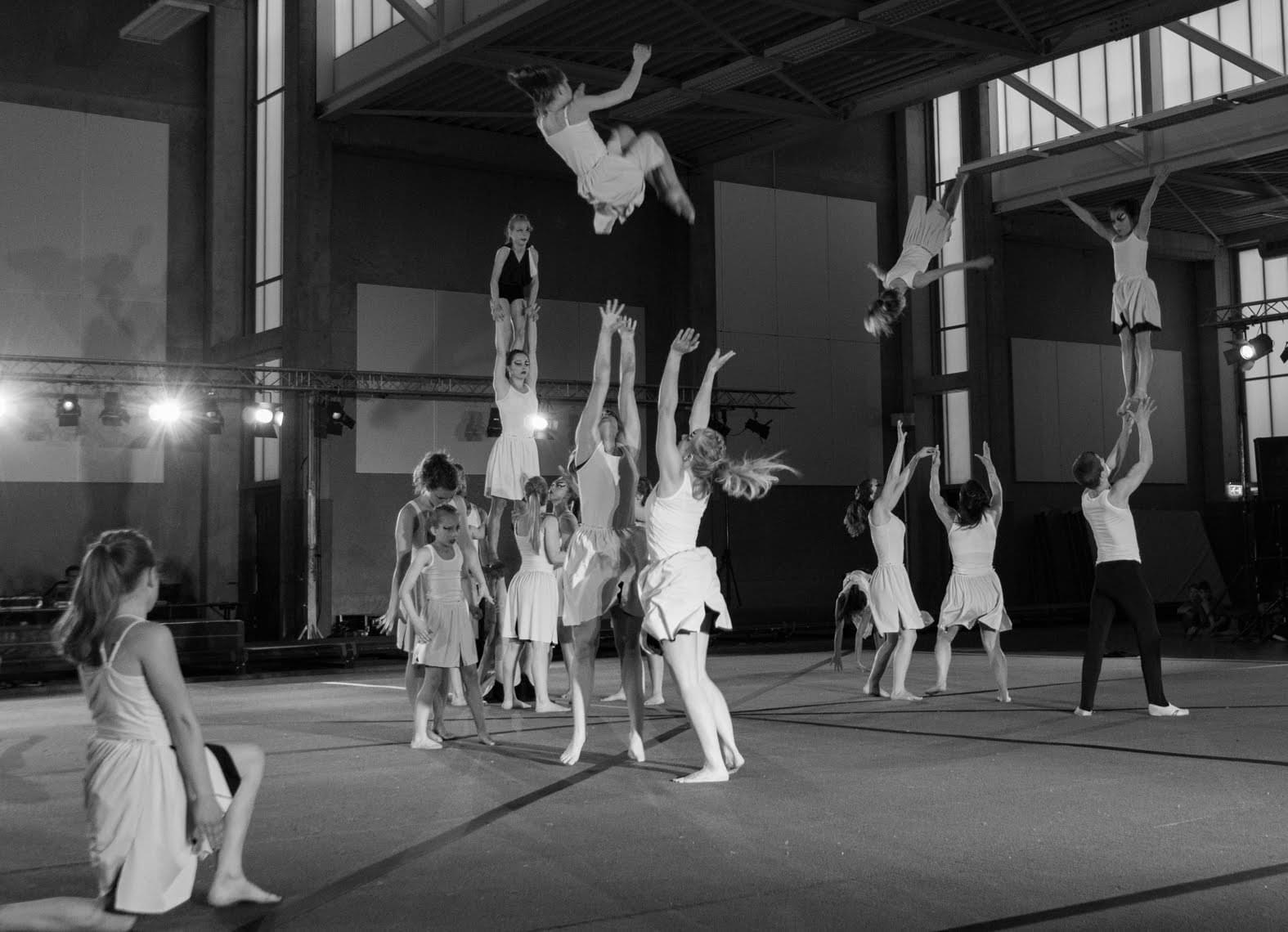 Turners van Turnclub Olympia Zwijnaarde voeren een indrukwekkende acrobatische show uit, waarbij ze in de lucht worden gegooid en spectaculaire sprongen maken. De atleten, gekleed in witte outfits, tonen hun kracht en coördinatie tijdens deze synchronische uitvoering in de turnhal van de club.