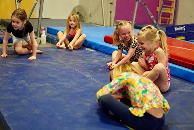 Kleuterturnen bij Turnclub Olympia Zwijnaarde - een groep jonge meisjes geniet van een speelse gymnastieksessie op de turnmat. De kinderen lachen en hebben plezier terwijl ze verschillende oefeningen uitvoeren. Ideaal voor jonge kinderen om hun motorische vaardigheden te ontwikkelen en nieuwe vriendjes te maken.