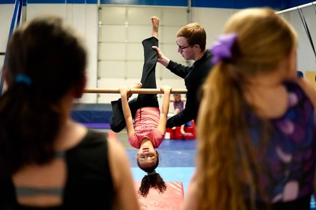 Jeugdgymnastiek training bij Turnclub Olympia Zwijnaarde - een meisje oefent een oefening aan de brug met hulp van een trainer, terwijl andere kinderen toekijken. Perfecte omgeving voor kinderen om gymnastiekvaardigheden te leren en zelfvertrouwen op te bouwen.