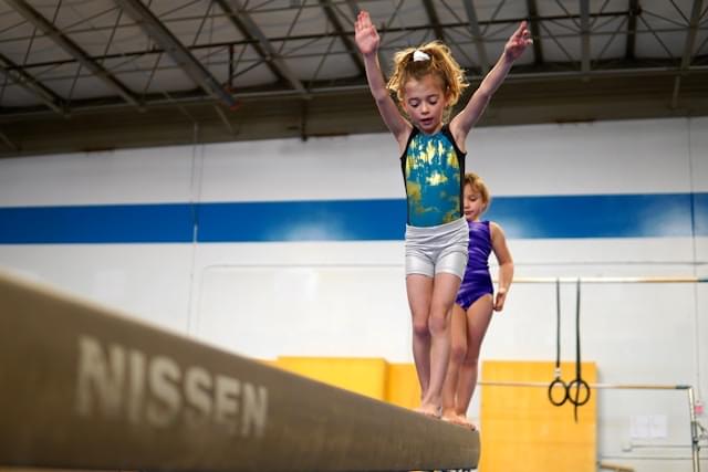 Twee jonge gymnasten oefenen op de balk in de turnzaal van Turnclub Olympia Zwijnaarde. De voorste gymnast balanceert op de balk met de armen omhoog. De turnzaal is uitgerust met professioneel trainingsmateriaal.