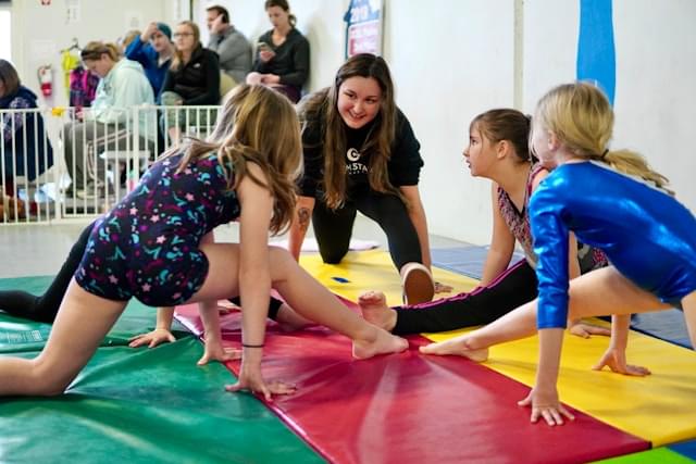 Gymnastiektraining bij Turnclub Olympia Zwijnaarde - meisjes in kleurrijke turnkleding doen stretch oefeningen onder begeleiding van een coach. Ouders kijken toe terwijl hun kinderen genieten van een leerzame en plezierige training.